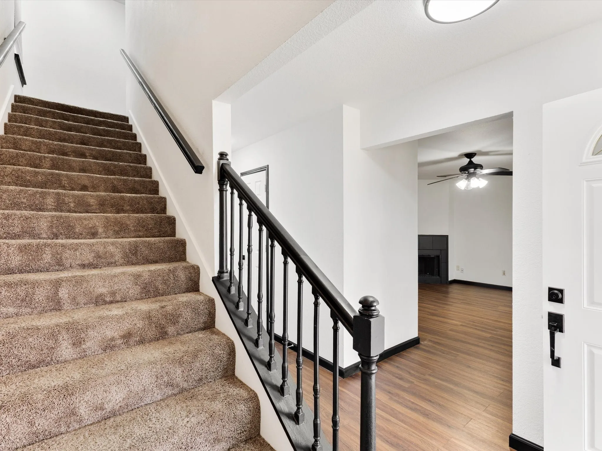 Carpeted staircase with a black wood banister, white walls, wood-finish flooring, a black fireplace, and a ceiling fan with integrated lighting