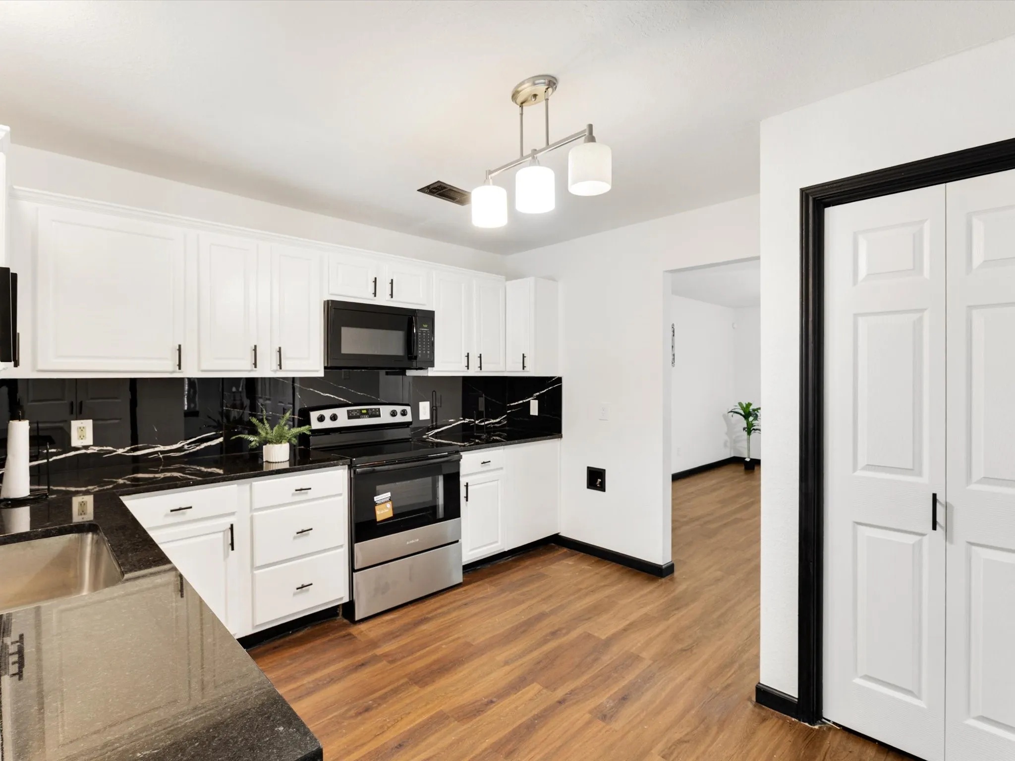 White shaker-style cabinetry with black hardware, black marble-look countertops and backsplash, stainless steel appliances, wood-finish flooring, and a contemporary ceiling light fixture