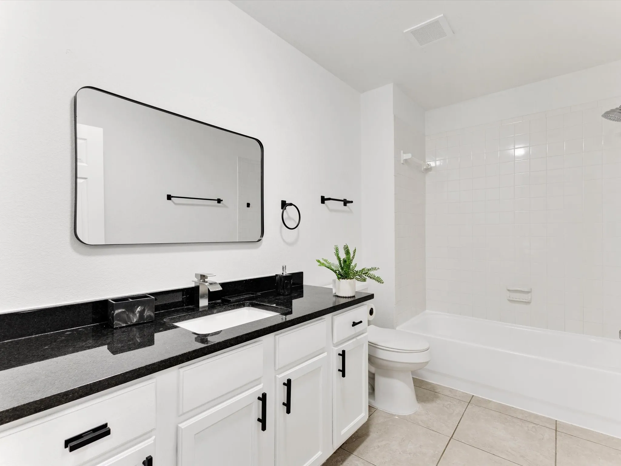 White cabinetry with black hardware, black countertop, rectangular undermount sink, wall-mounted mirror, and a white tiled shower-tub combination