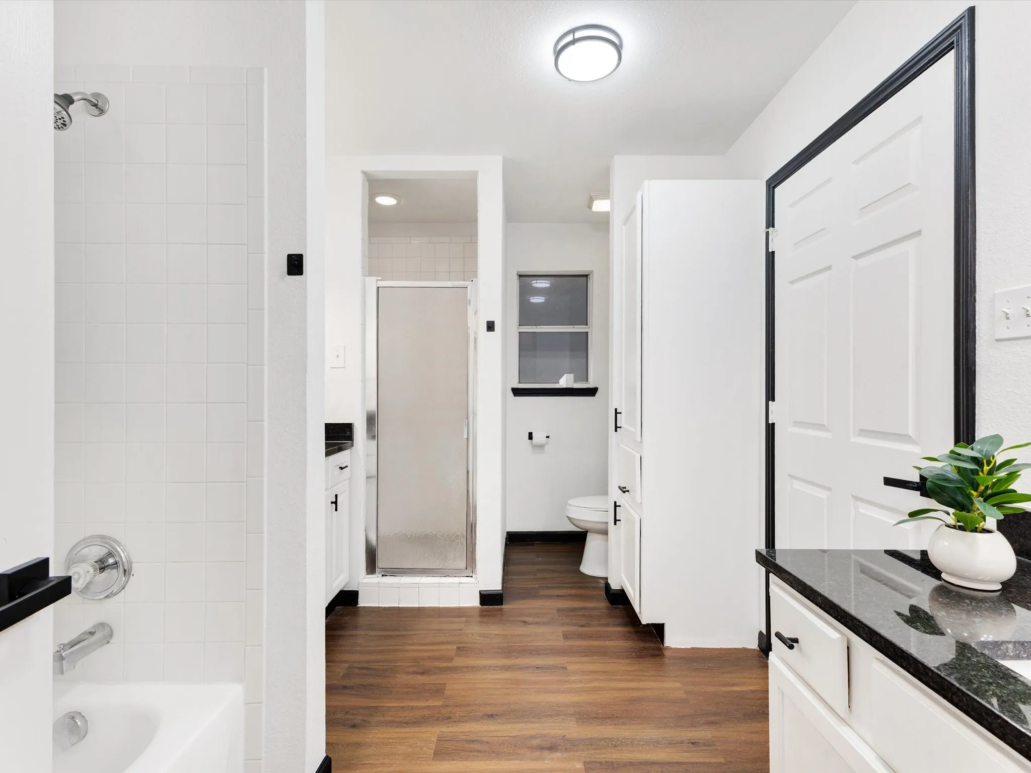 Bathroom featuring wood-finish flooring, white cabinetry, black countertops, and a combination tub/shower with white tile surround