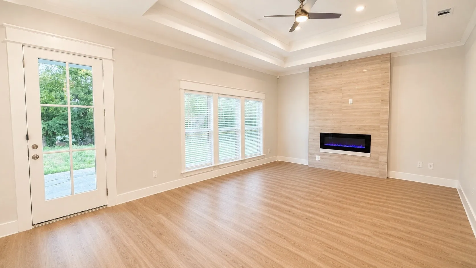 Spacious living area featuring wood-finish flooring, a contemporary fireplace with a tiled surround, a multi-tiered tray ceiling with a ceiling fan, and ample natural light from multiple windows