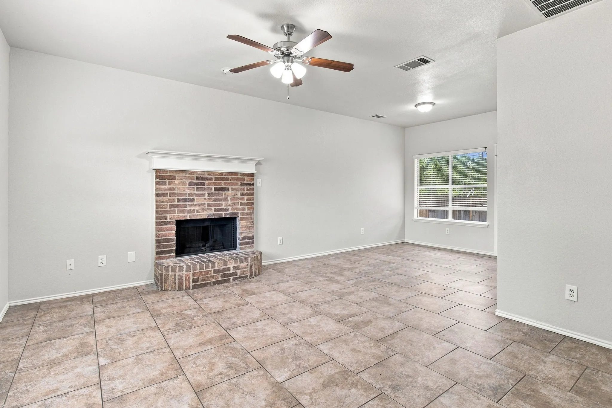 Spacious living area featuring a brick fireplace, tiled flooring, ceiling fan, and ample natural light from the window