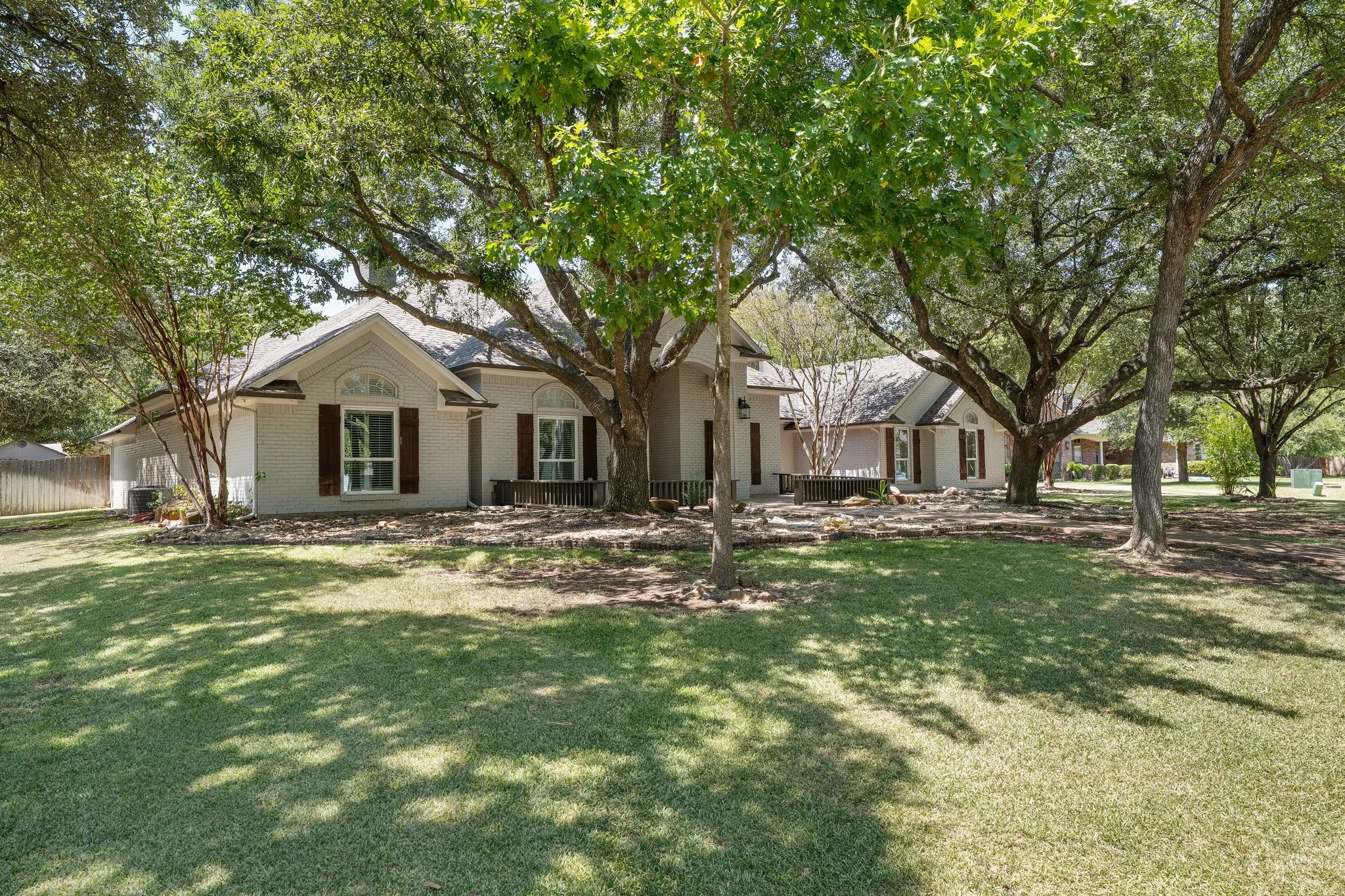 Spacious front yard with mature trees, stone landscaping, and a white exterior home with dark shutters