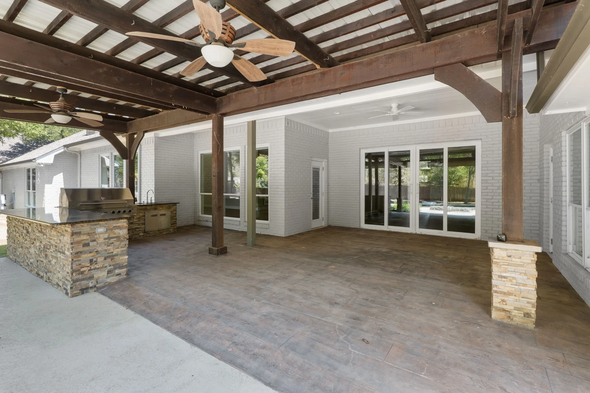 Covered patio featuring an outdoor kitchen with built-in grill, stone veneer accents, and a wood-beamed ceiling with ceiling fans
