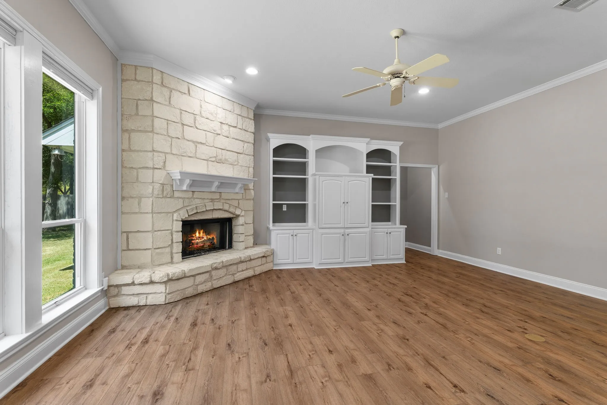 Corner stone fireplace with hearth, built-in cabinetry, wood-finish flooring, and ceiling fan