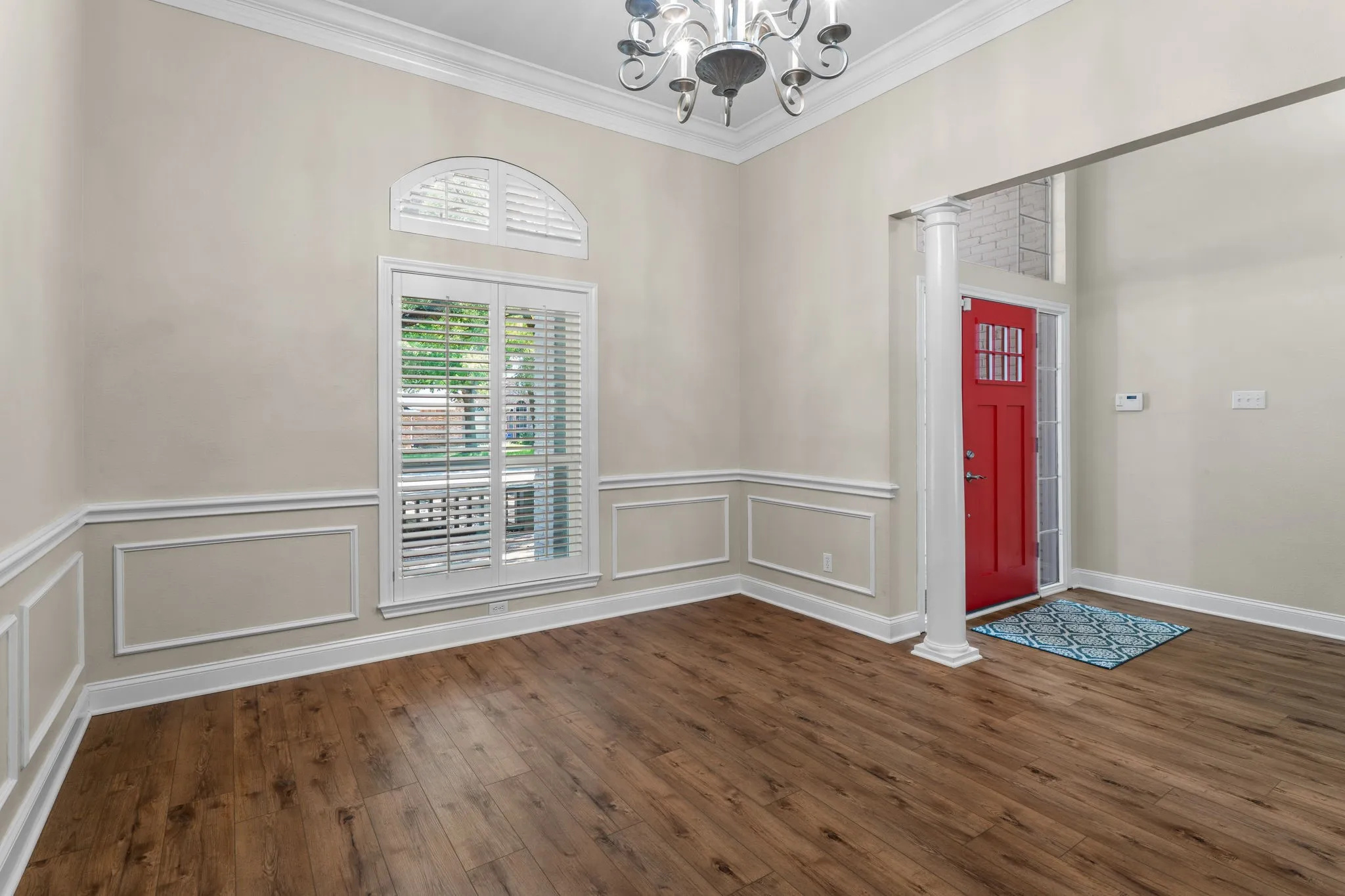 Wood-finish flooring, wainscoting, crown molding, arch window, and a red paneled entry door