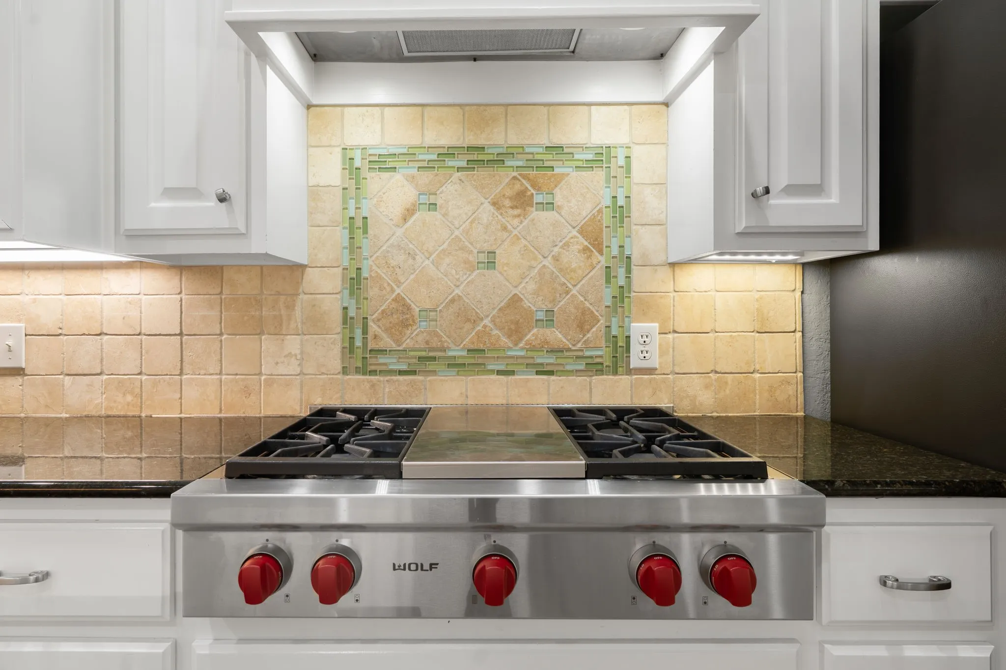 Stainless steel range, stone tile backsplash with decorative inset, granite countertops, white cabinetry with silver hardware, and under-cabinet lighting