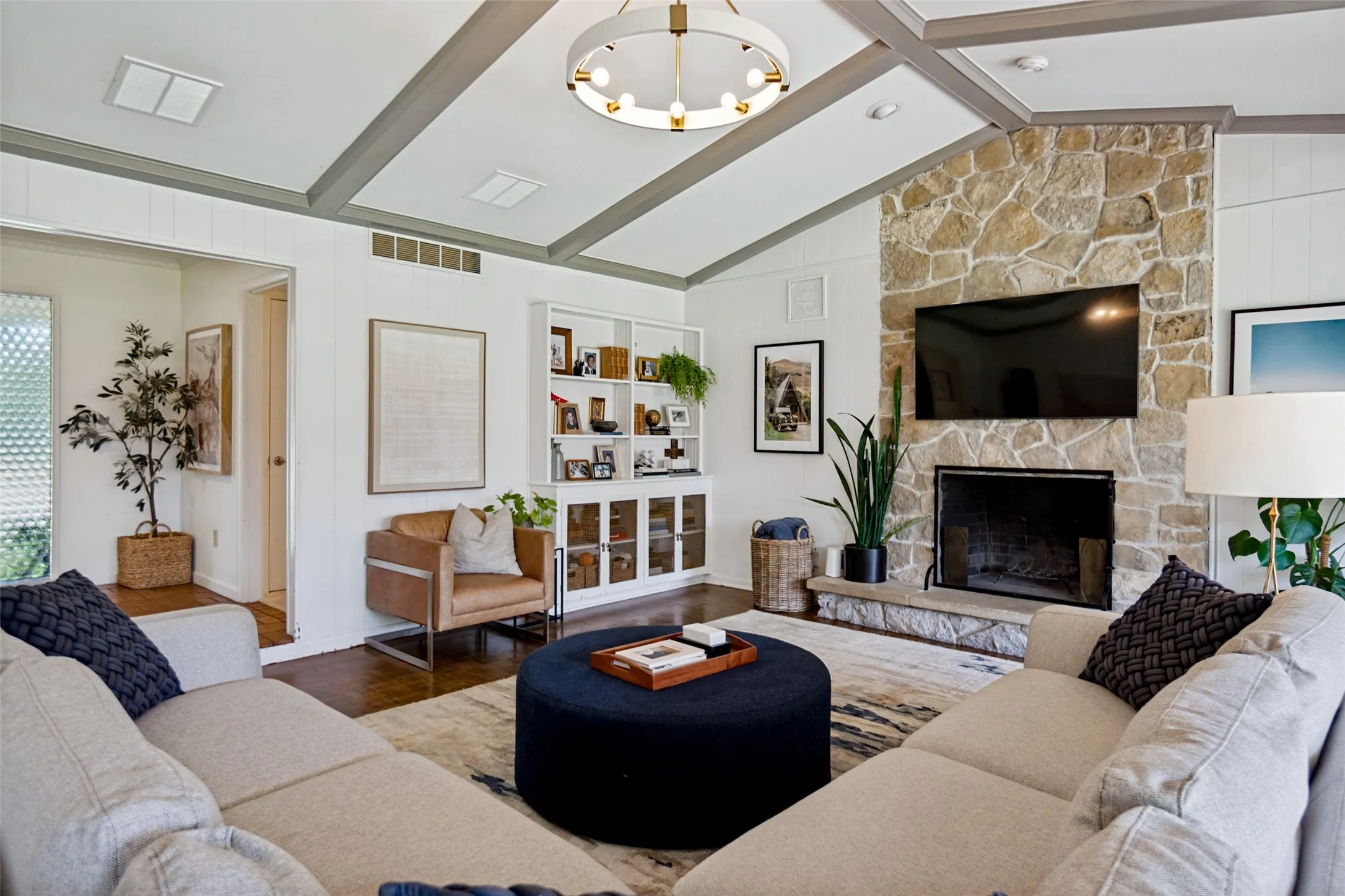 Vaulted ceiling with exposed beams, stone-clad fireplace, built-in shelving, wood-finish flooring, and a modern chandelier