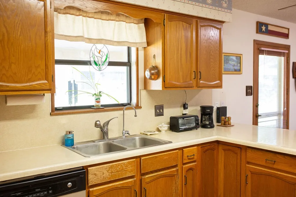 Wood-finish cabinetry, double basin stainless steel sink, light-toned countertops, and a window with a stained glass accent