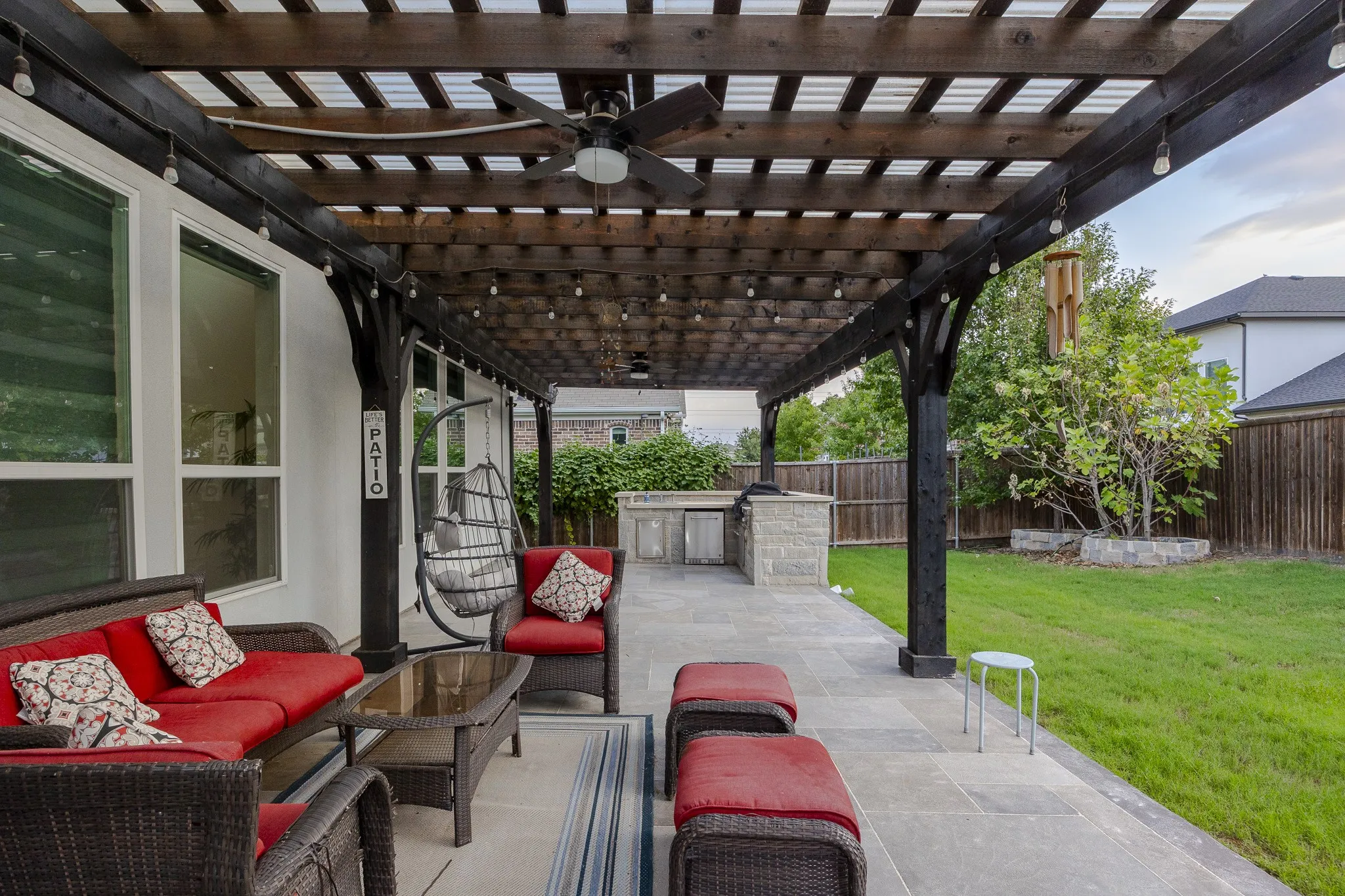 Expansive covered patio featuring a wood pergola with integrated lighting, a ceiling fan, a built-in outdoor kitchen with stone detailing, and a spacious green lawn