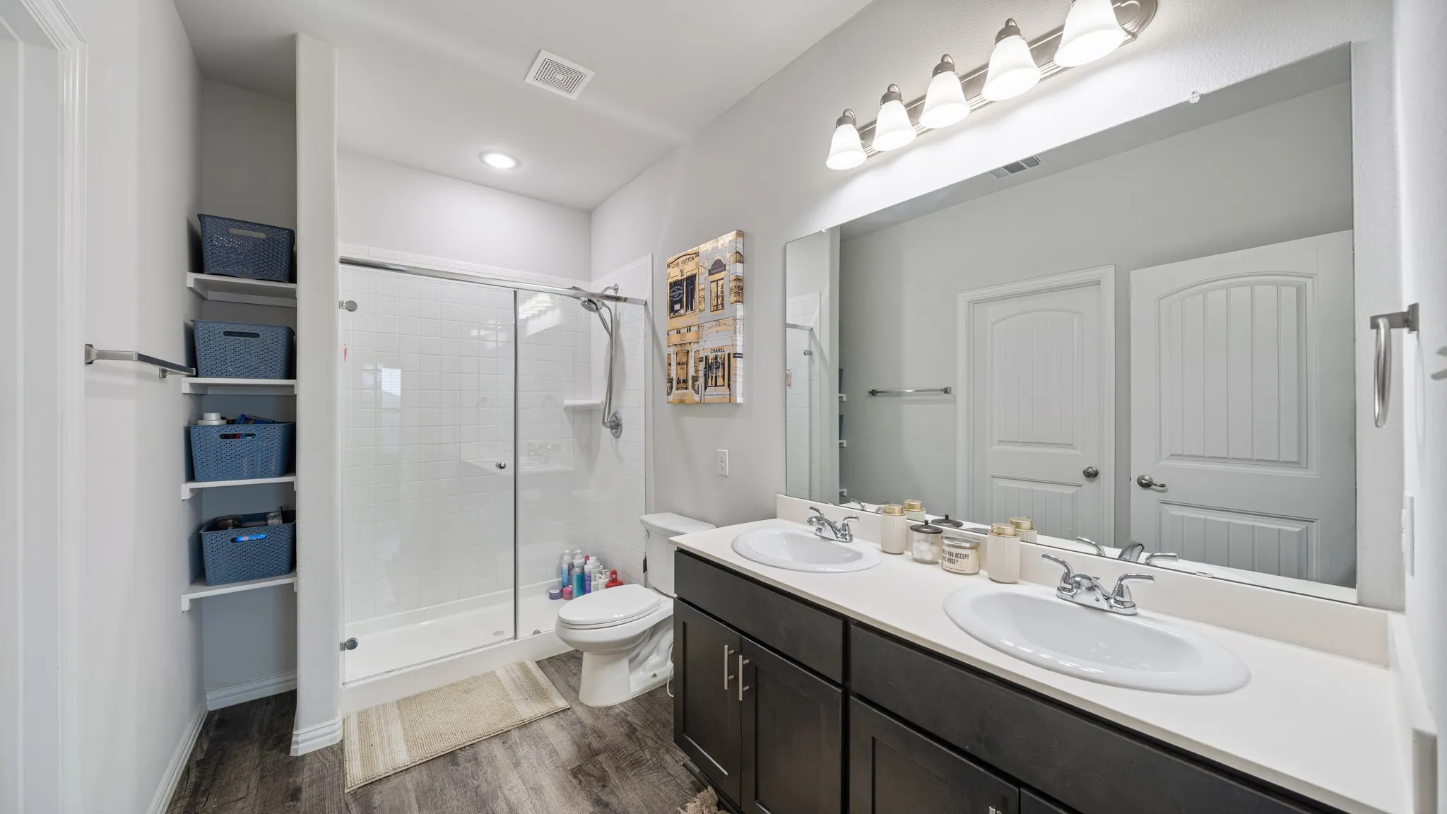 Dual vanity with white countertop and dark cabinetry, large frameless mirror, glass-enclosed shower, built-in shelving, and wood-finish flooring