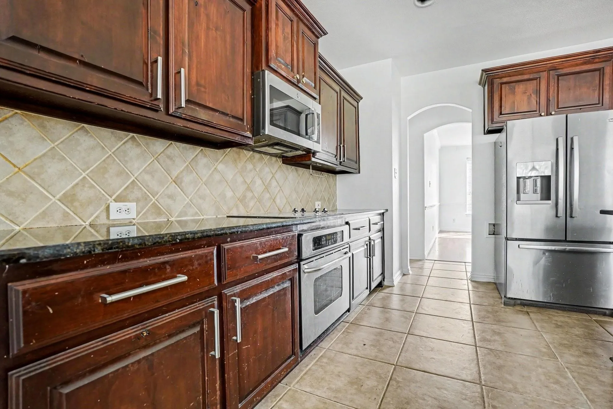 Dark wood-finish cabinetry with brushed nickel hardware, built-in stainless steel appliances, dark stone countertops, tile backsplash, and tile flooring