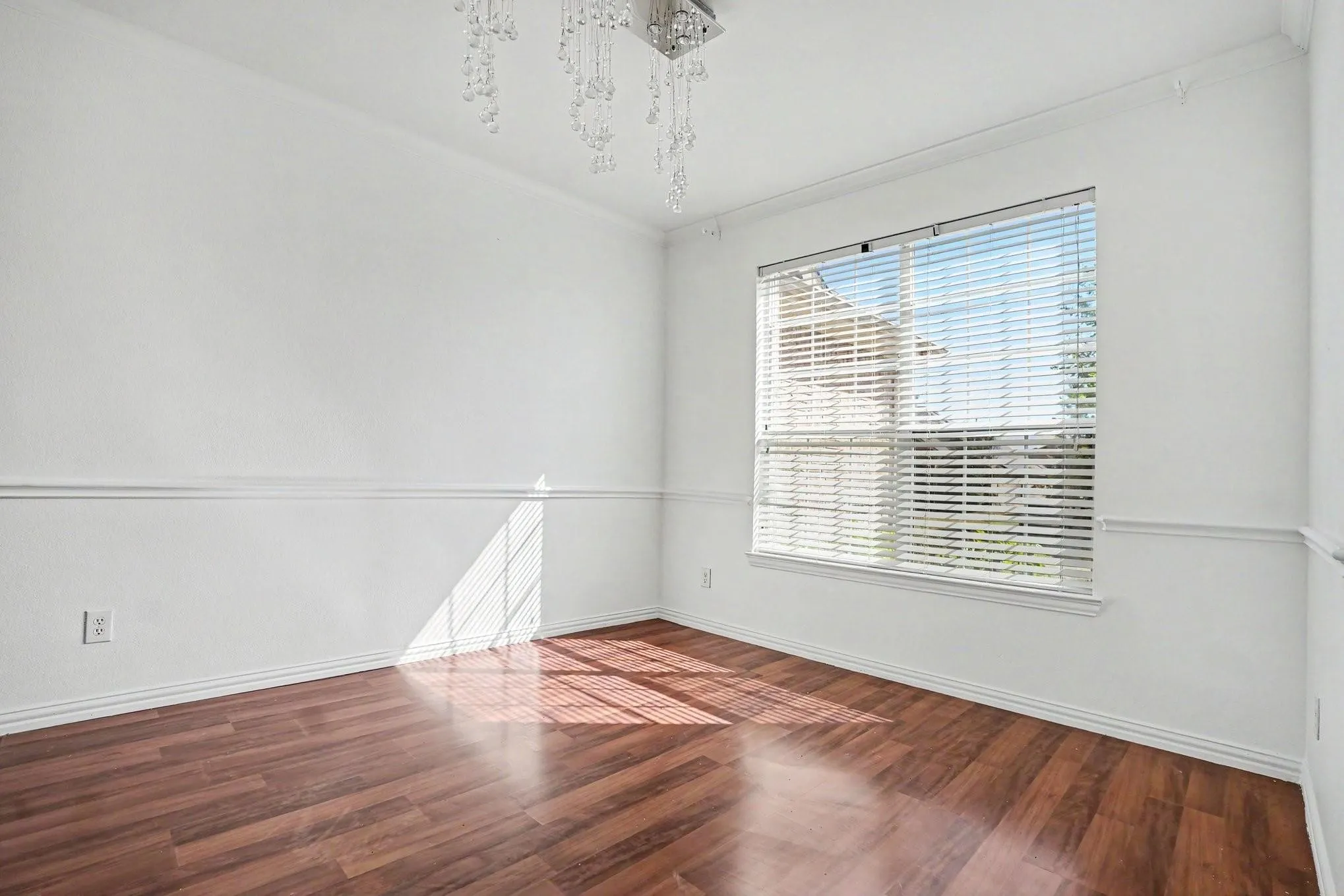 Hardwood-style flooring, white walls with decorative chair rail molding, a large window with horizontal blinds, and a multi-tiered crystal chandelier