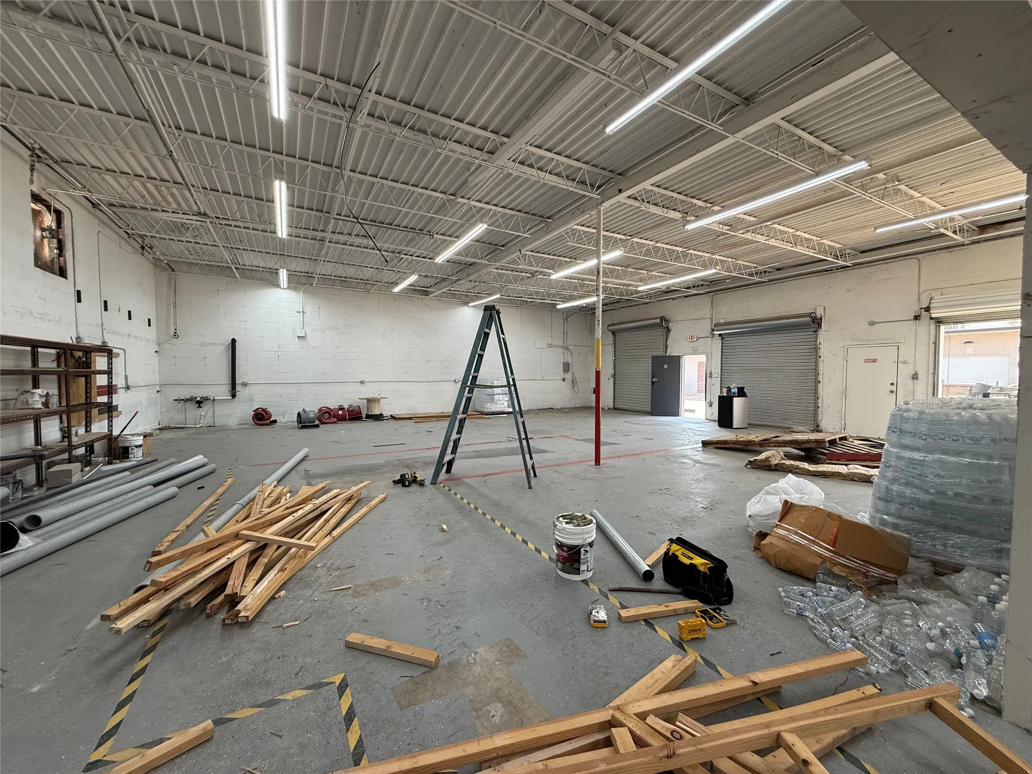 Spacious commercial interior featuring a high trussed ceiling, multiple fluorescent light fixtures, two roll-up doors, and an additional personnel door