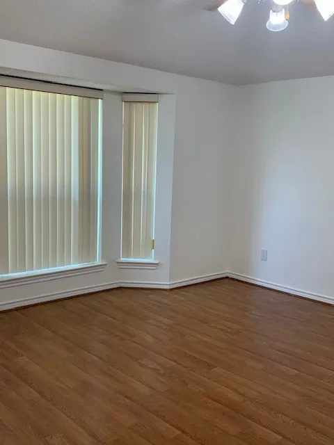 Wood-finish flooring throughout, bay window with vertical blinds, and a ceiling fan with integrated lighting