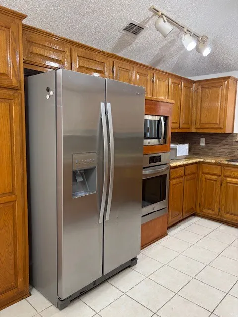 Stainless steel side-by-side refrigerator, built-in stainless steel microwave and oven, wood cabinetry, stone countertops, and tile flooring