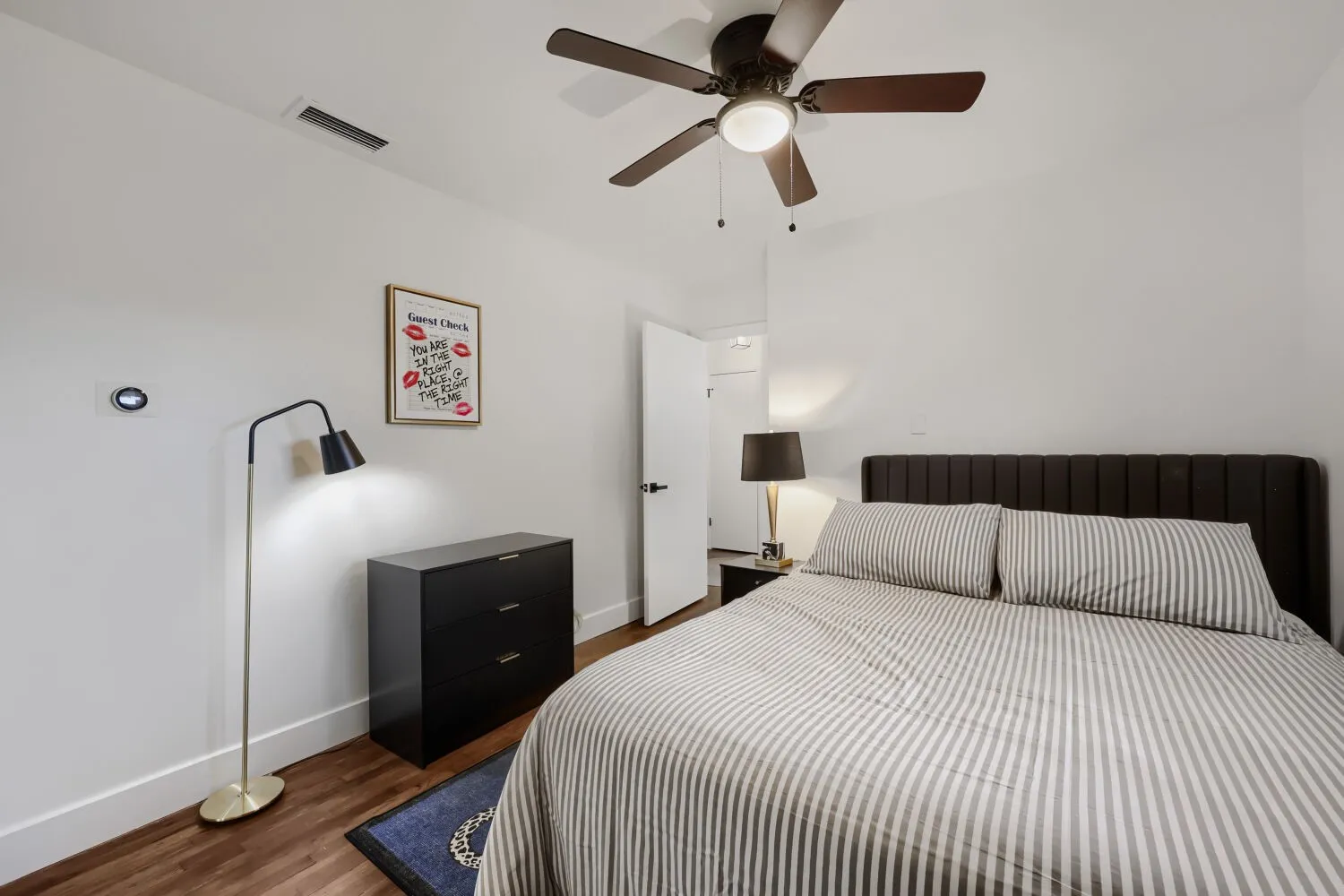 Ceiling fan with integrated lighting, wood-finish flooring, white walls, dark paneled headboard, and built-in wall thermostat