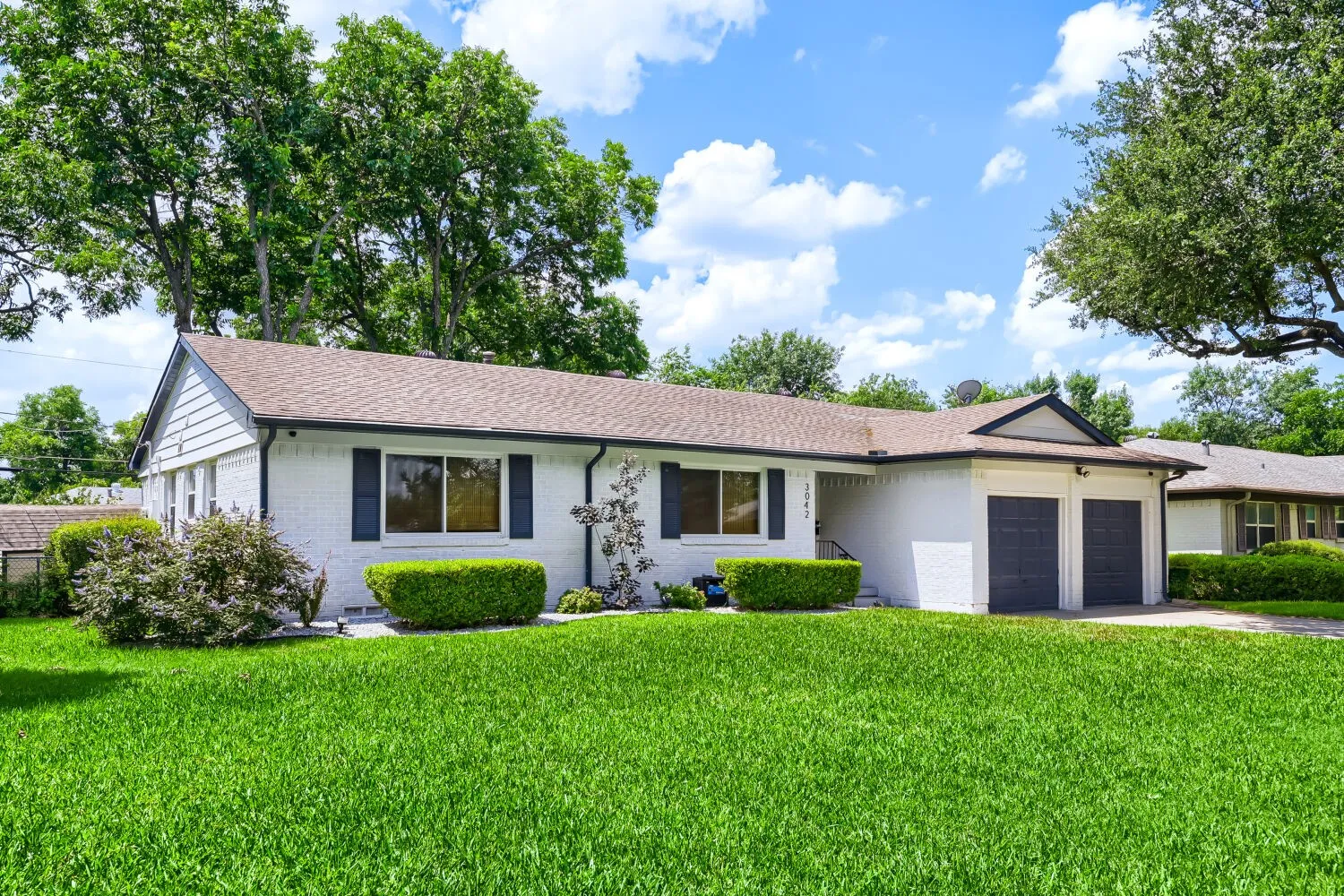 White brick exterior with dark shutters, two-car garage, and established landscaping