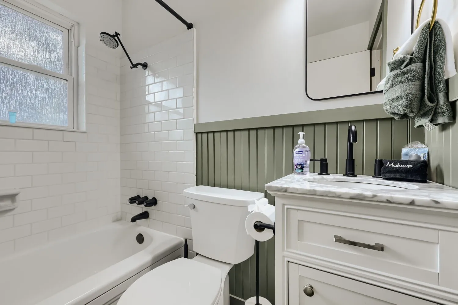 White subway tile shower surround, matte black fixtures, white vanity with stone-finish countertop, and decorative beadboard paneling