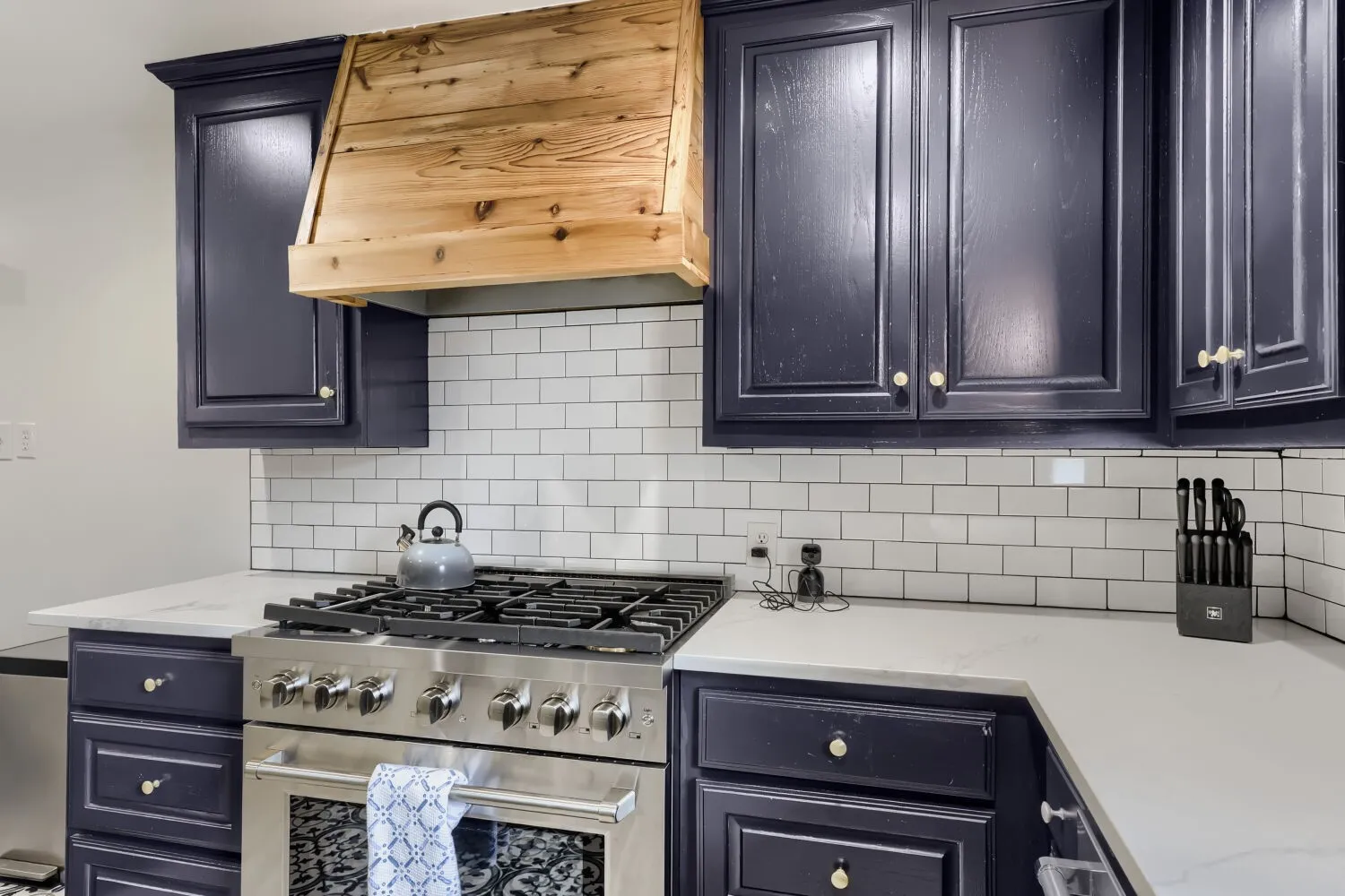 Stainless steel range, wood-finish range hood, white subway tile backsplash, dark-finish cabinetry, and light-finish countertops