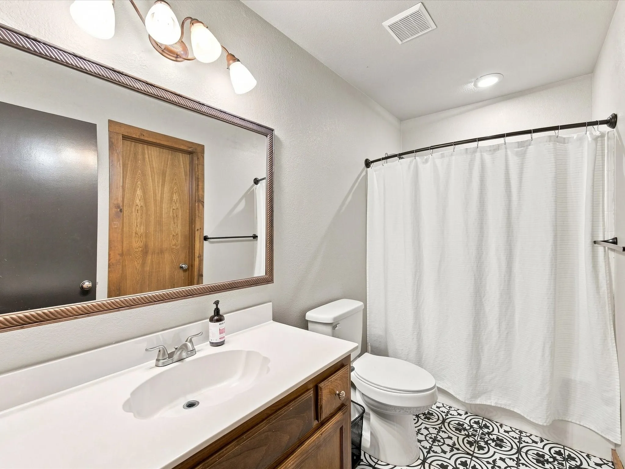 White countertop vanity with wood-finish cabinetry, decorative framed mirror, black and white patterned floor tiles, and a shower with a black curtain rod