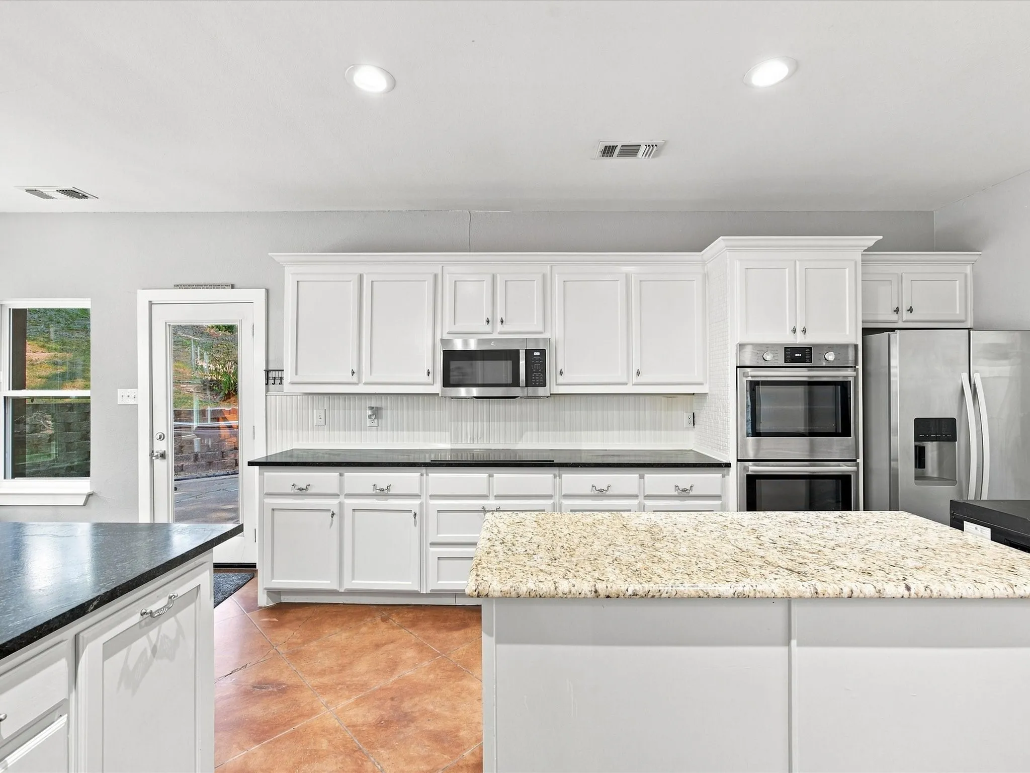 White cabinetry, stainless steel appliances, black countertops, island with light-colored countertop, and wood-finish flooring