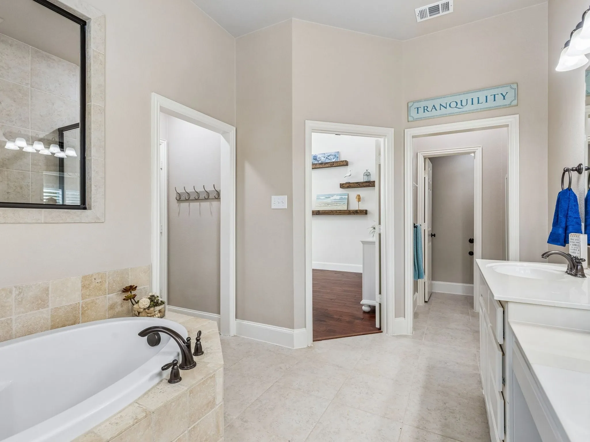 Soaking tub with stone surround, dual vanity with undermount sinks, bronze finish fixtures, and light-toned tile flooring.