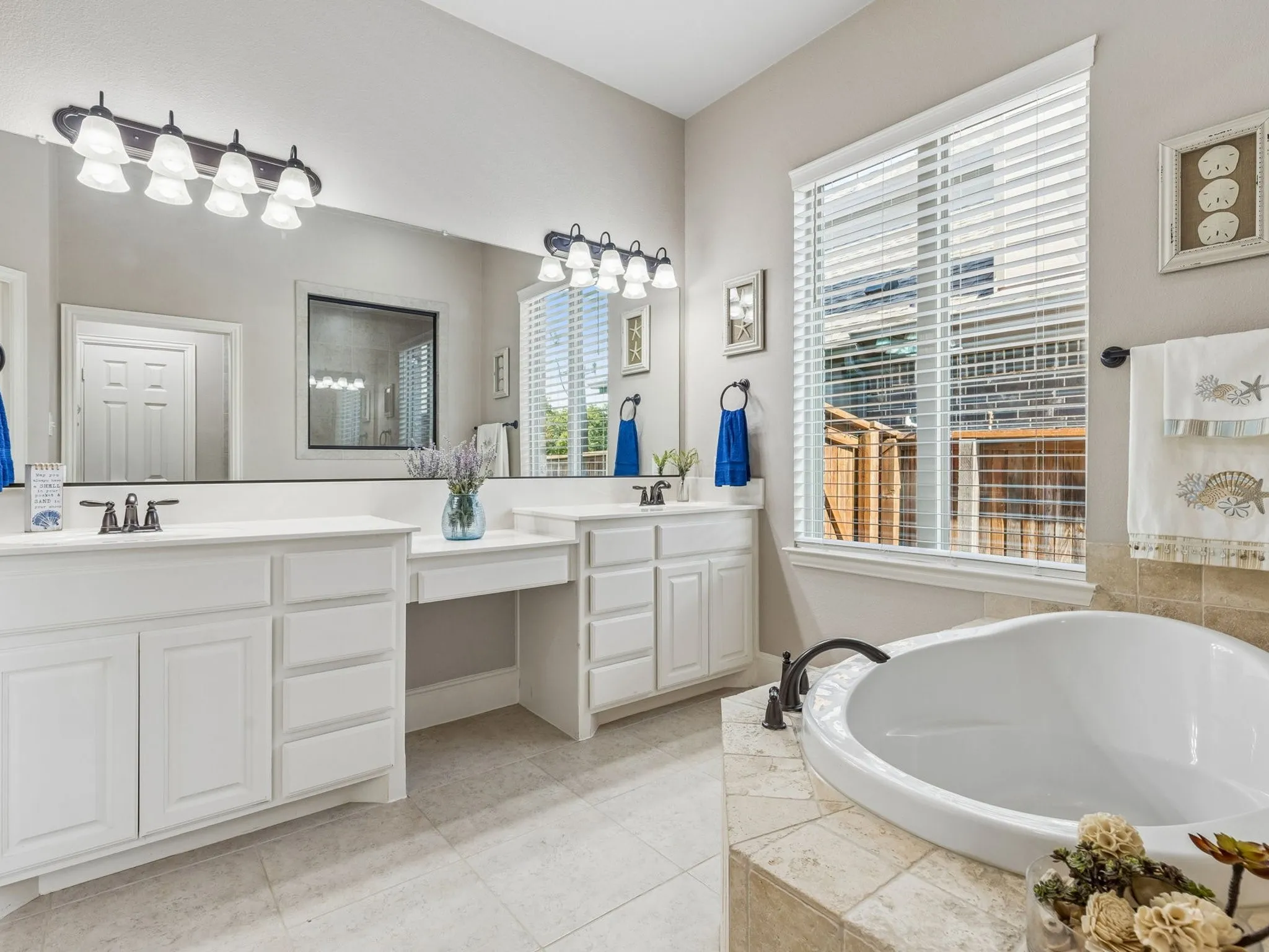 White dual vanities with integrated sinks, large vanity mirror, separate soaking tub, tiled tub surround, and light-toned tile flooring.