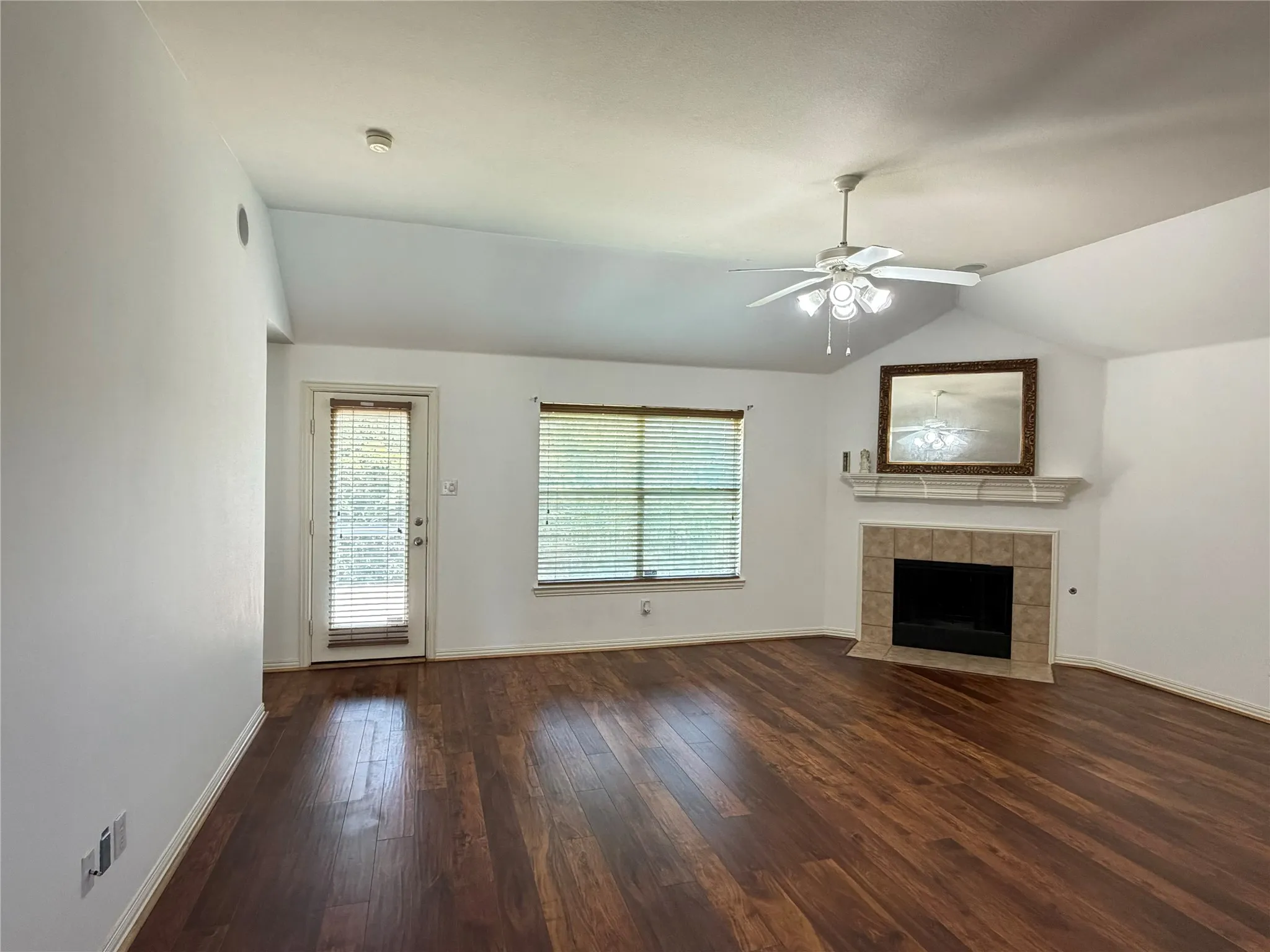 Vaulted ceiling with a ceiling fan, wood-finish flooring, fireplace with a tile surround and mantel, and a door with integrated blinds