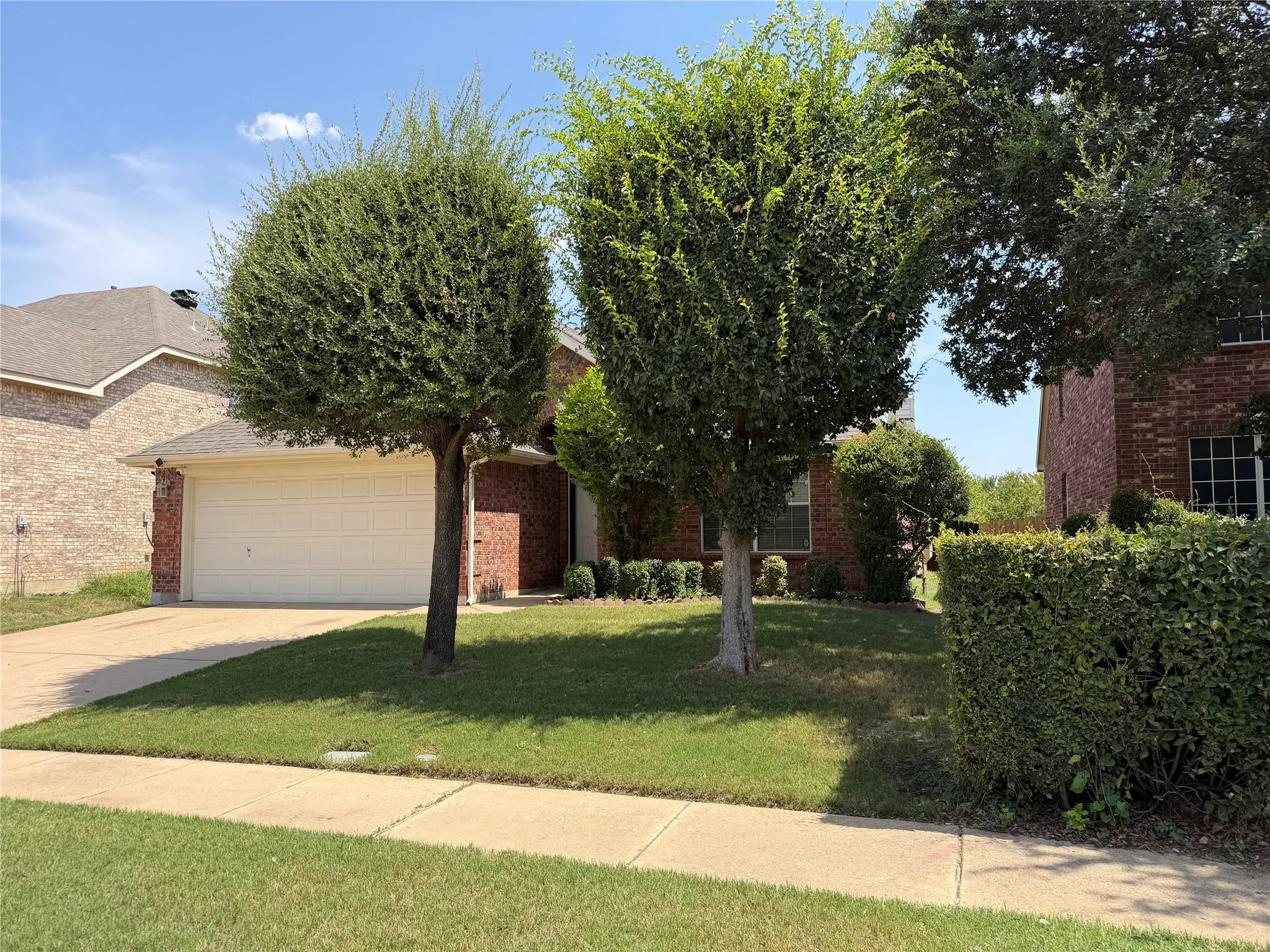 Brick exterior with an attached garage, paved driveway, and mature landscaping