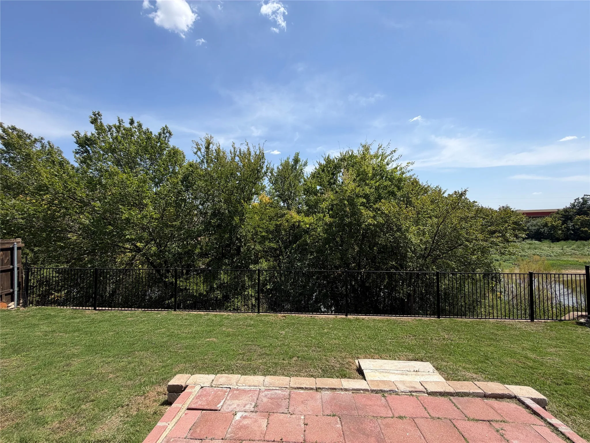 Expansive grass lawn with a paver patio and a black metal fence, bordered by mature trees and a body of water