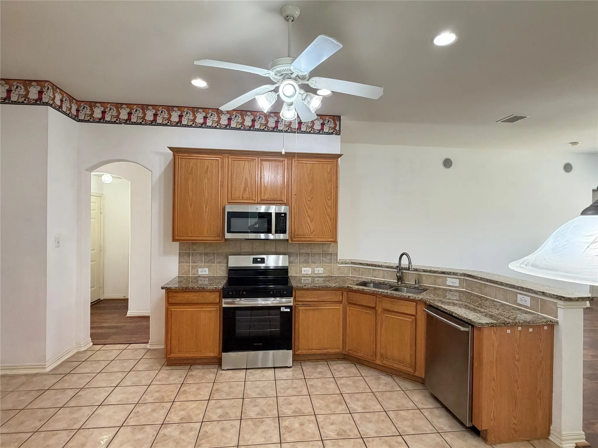 Kitchen featuring tile flooring, wood cabinetry, stainless steel appliances, granite countertops, and a ceiling fan with integrated lighting