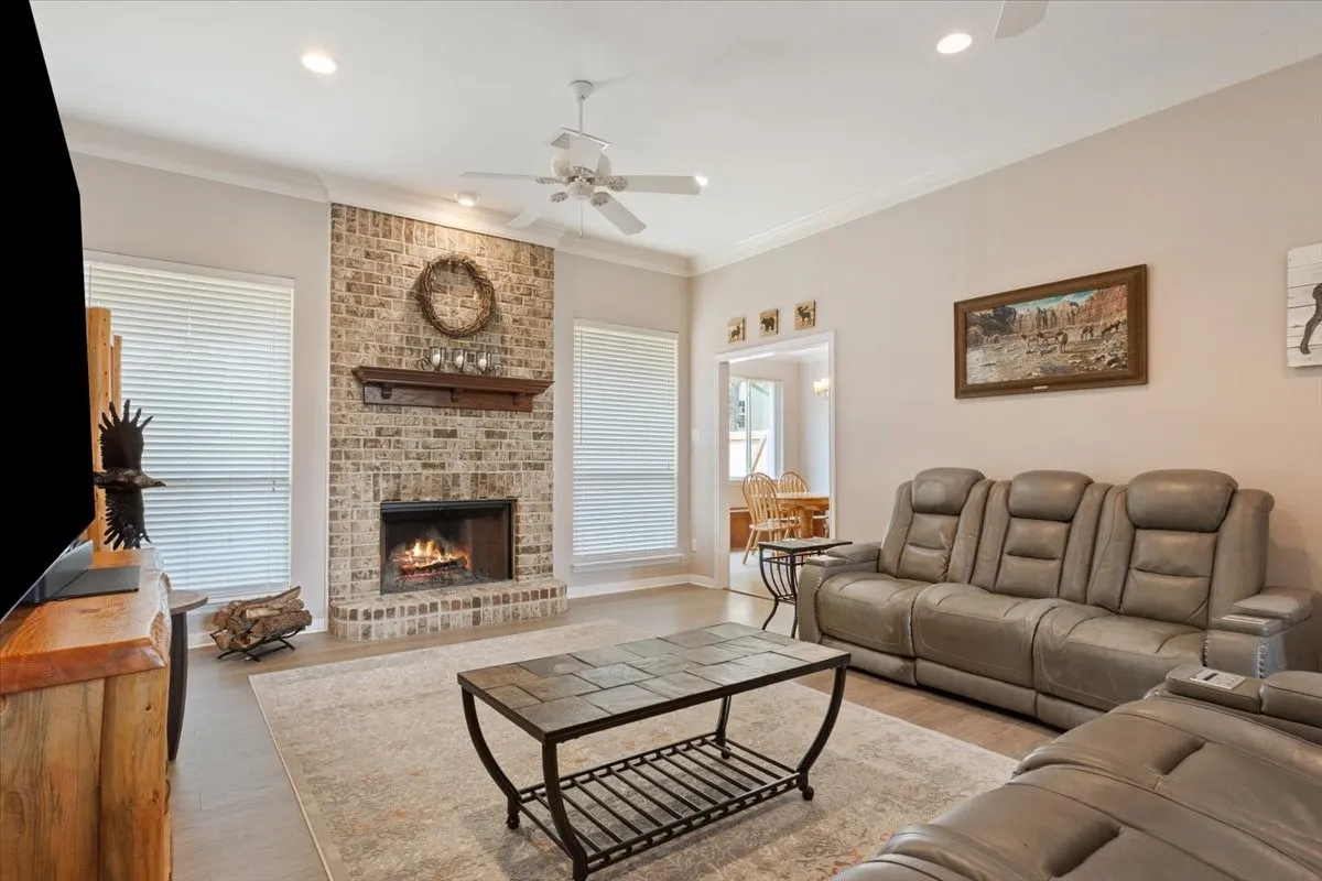 Spacious living area featuring a brick-clad fireplace with a wood mantel, crown molding, a ceiling fan, and recessed lighting