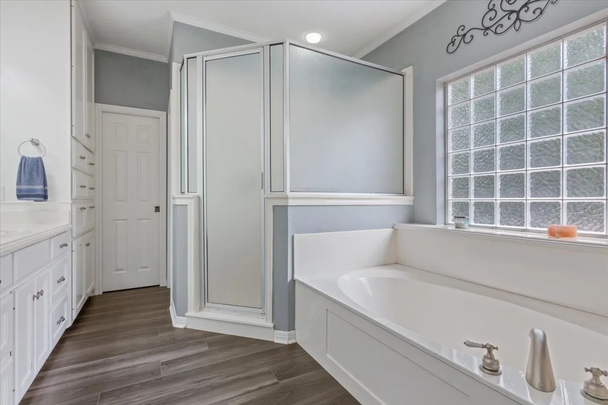 Corner shower with frosted glass enclosure, soaking tub with brushed nickel fixtures, glass block window, double vanity with white cabinetry, and wood-finish flooring