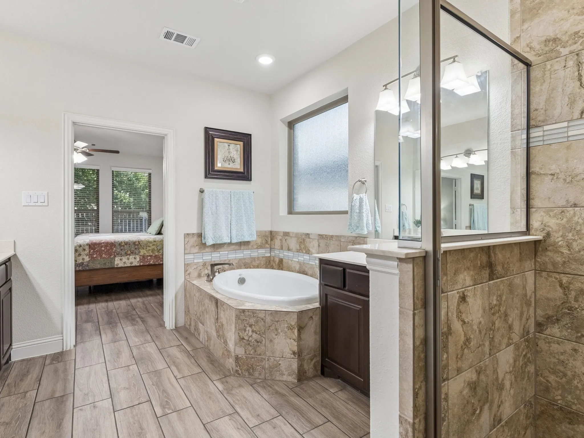 Corner soaking tub with tile surround, wood-finish tile flooring, and a frosted privacy window