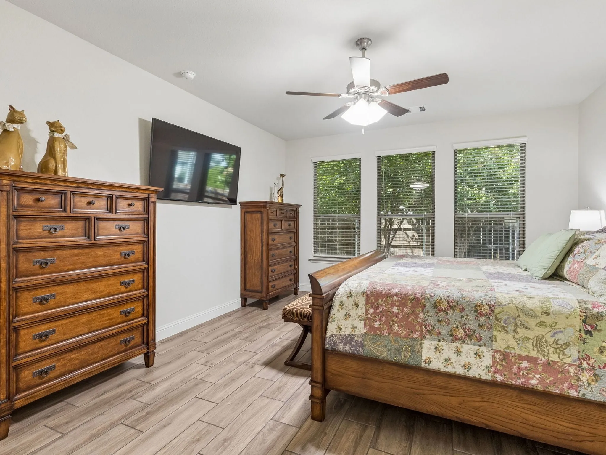 Master bedroom featuring wood-finish tile flooring, ceiling fan with integrated lighting, a wall-mounted flat-screen television, and abundant natural light from multiple glass doors and windows overlooking the surrounding landscape