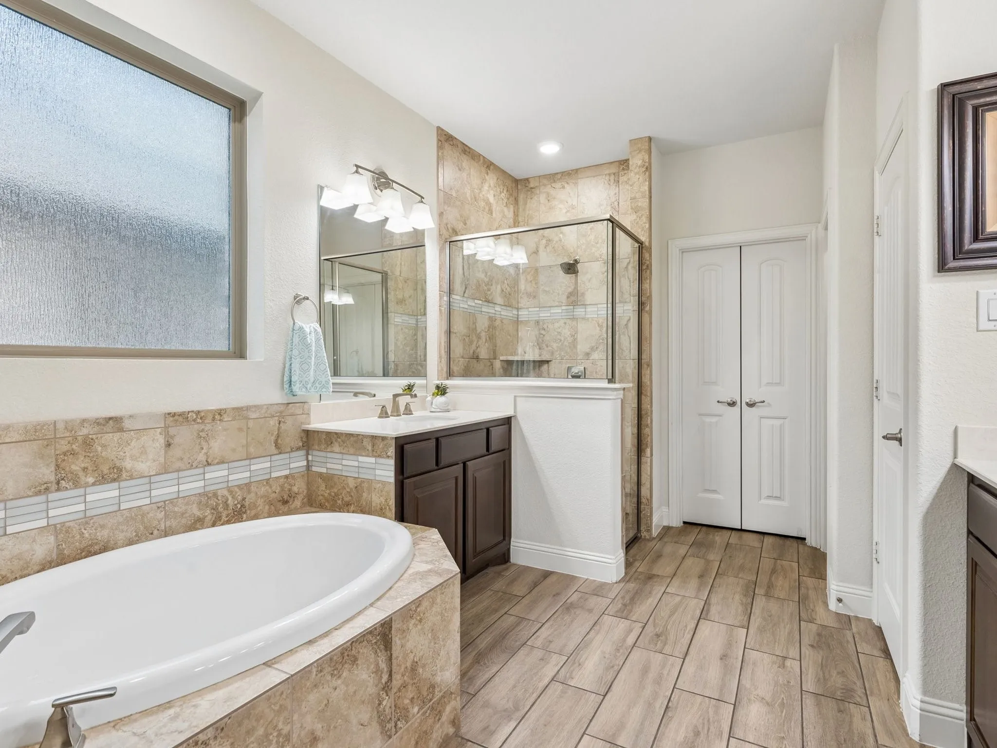 Soaking tub with tiled surround, wood-finish tile flooring, double-door entry, and a glass-enclosed shower