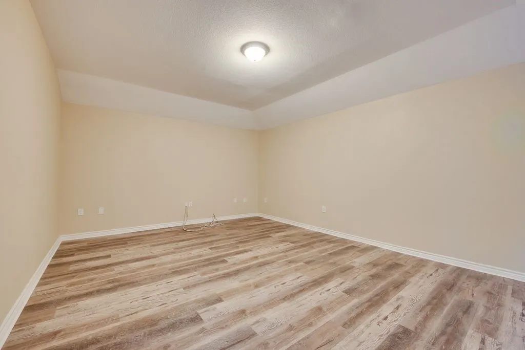 Spacious room featuring wood-finish flooring, light neutral walls, and a tray ceiling with a central flush-mount light fixture