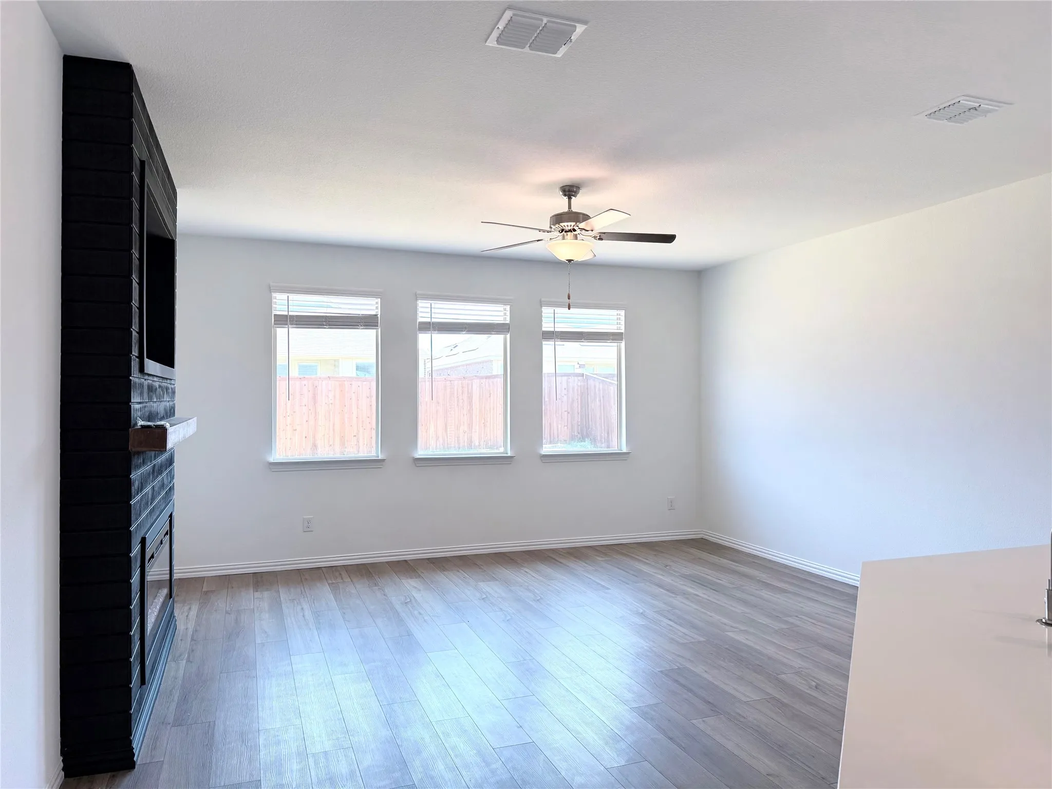 Wood-finish flooring, white walls, black brick-style fireplace surround with wooden mantel, three windows, and a ceiling fan with integrated lighting