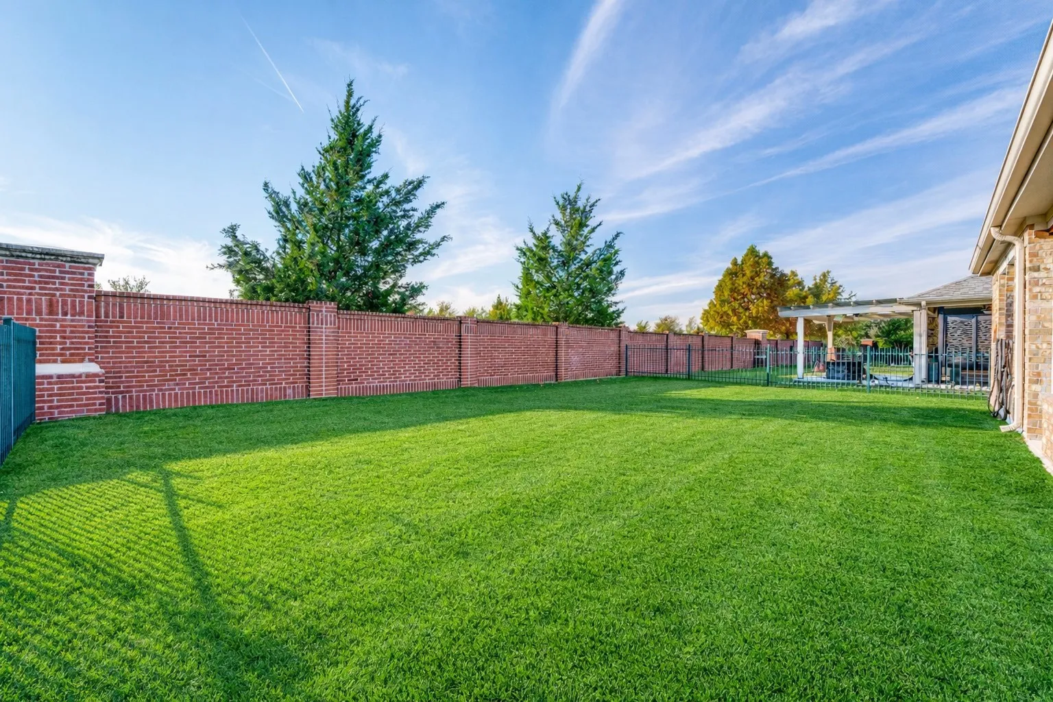 Expansive backyard with a manicured lawn, full brick privacy fencing, mature trees, and an outdoor patio with a pergola