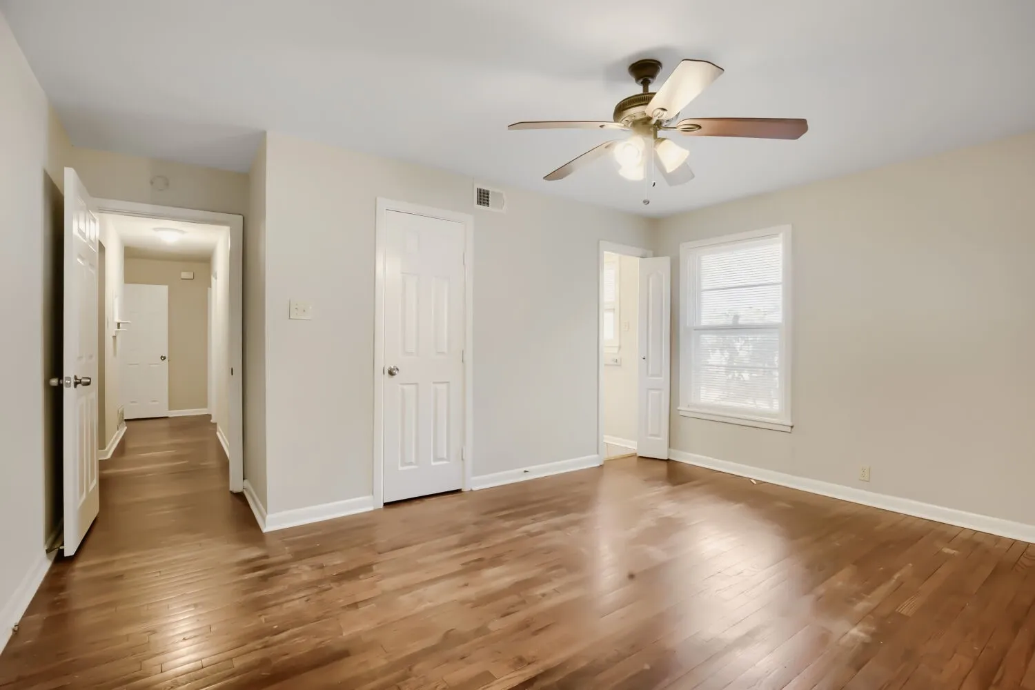 Spacious main living area showcasing gleaming wood-finish flooring throughout, abundant natural light from large windows, ceiling fan with integrated lighting, and multiple white six-panel doors providing access to adjoining spaces
