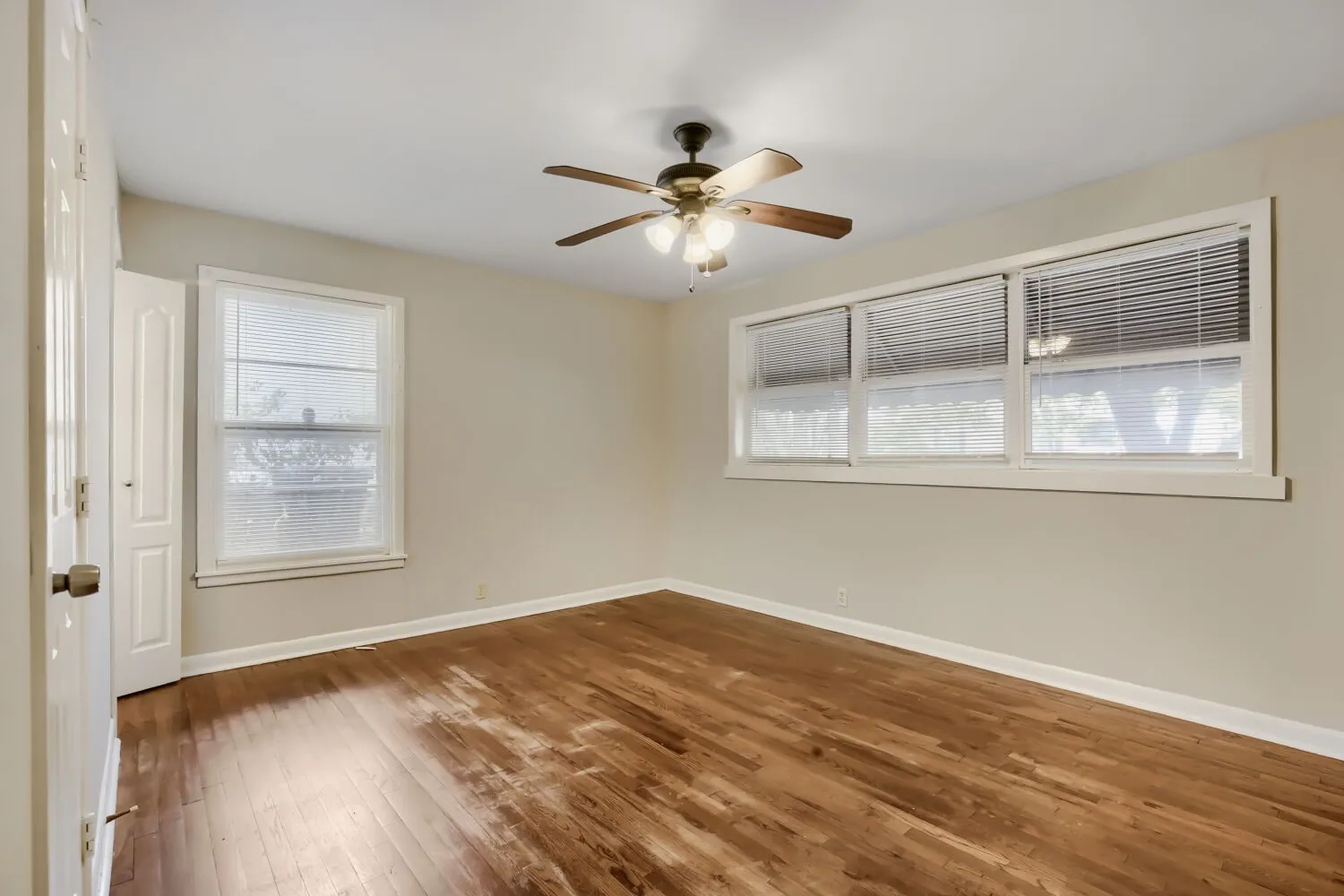 Bright corner room showcasing rich wood-finish flooring, ceiling fan with integrated lighting, and multiple windows providing abundant natural light