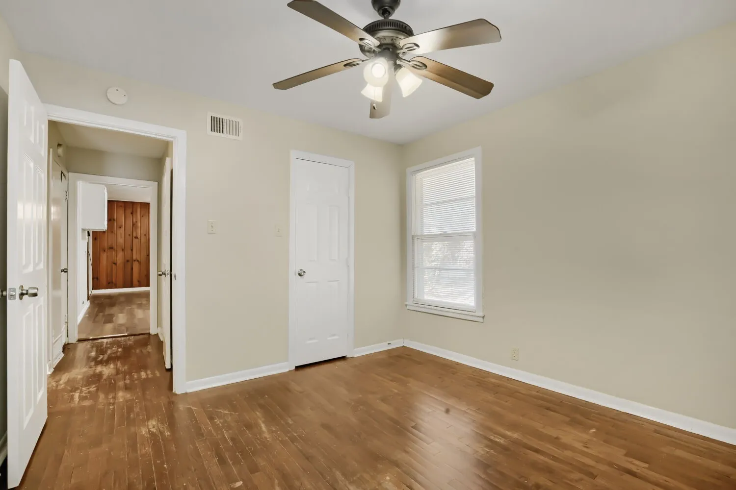 Spacious bedroom with warm wood-finish flooring, ceiling fan with integrated lighting, large window with natural light, and white trim throughout