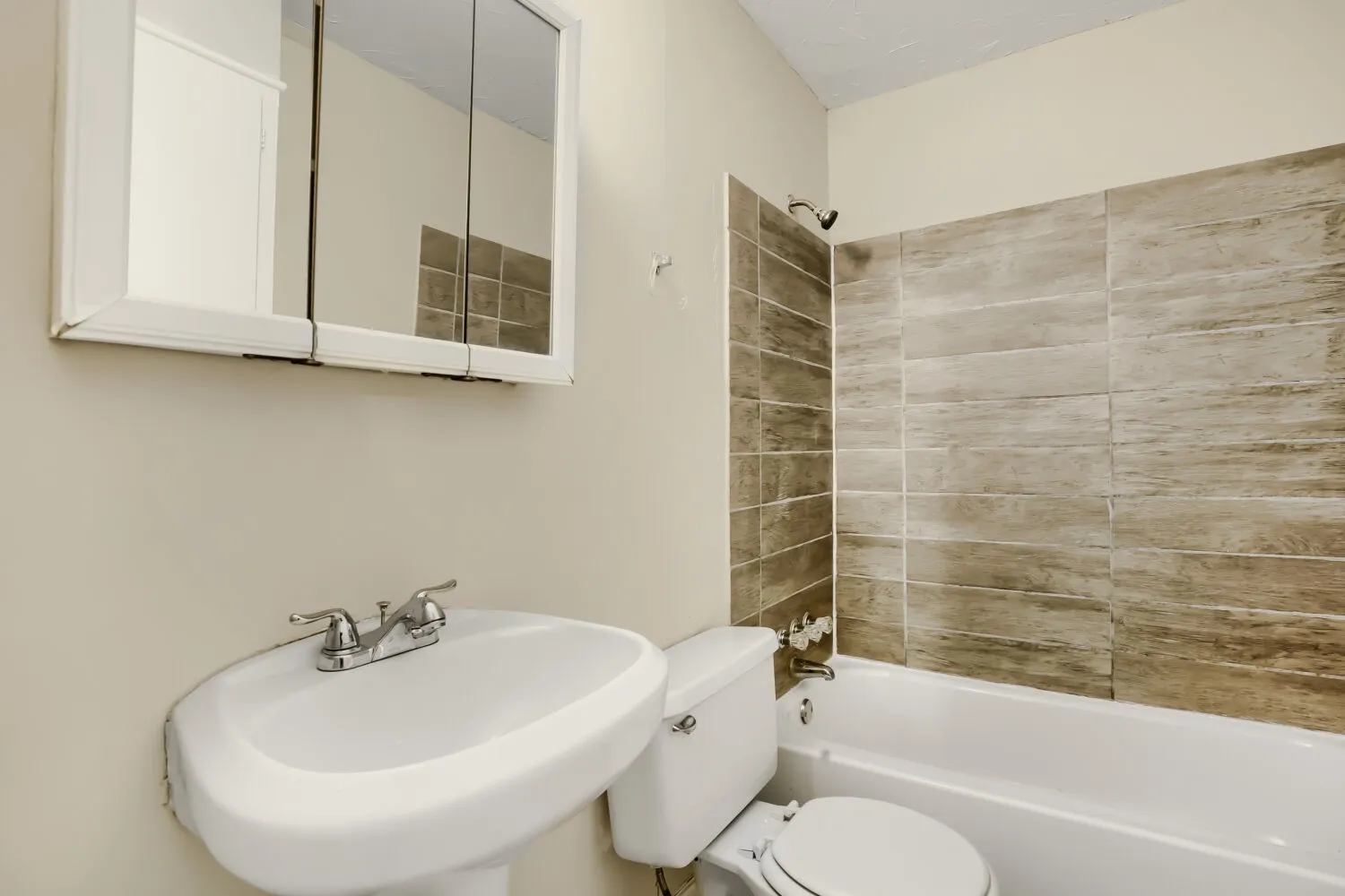 Bright bathroom featuring a curved pedestal sink with chrome faucet, mirrored medicine cabinet, wood-finish tile shower surround with glass enclosure, and white subway tile accents