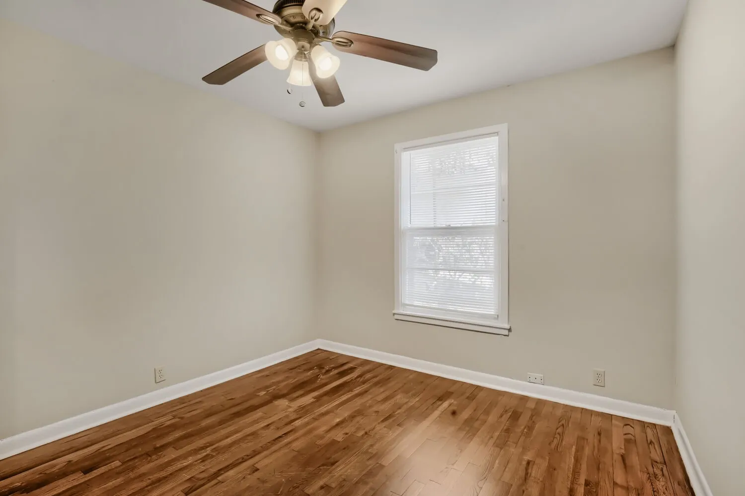 Bright corner bedroom showcasing gleaming wood-finish flooring, neutral walls, and abundant natural light from a large double-hung window