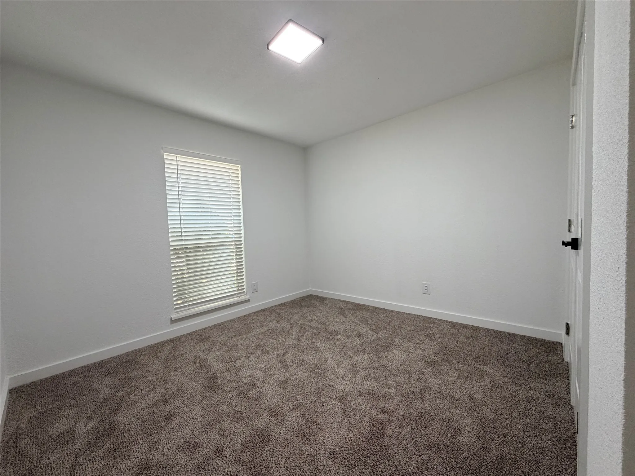 Neutral-toned carpet flooring, white painted walls, window with horizontal blinds, flush-mount ceiling light fixture, and a white interior door with a black handle