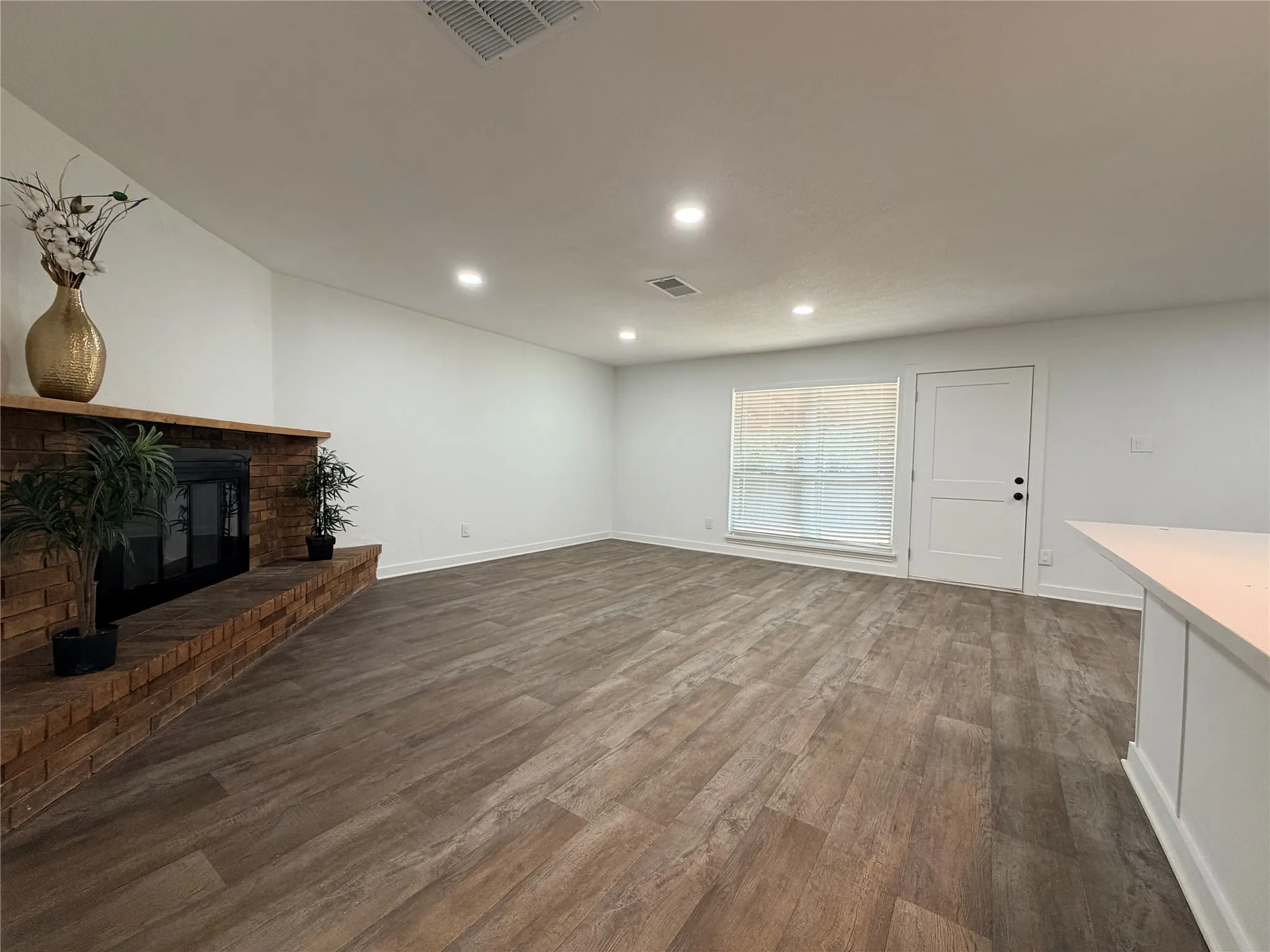 Spacious room with wood-finish flooring, recessed lighting, a brick fireplace, and a large window with blinds