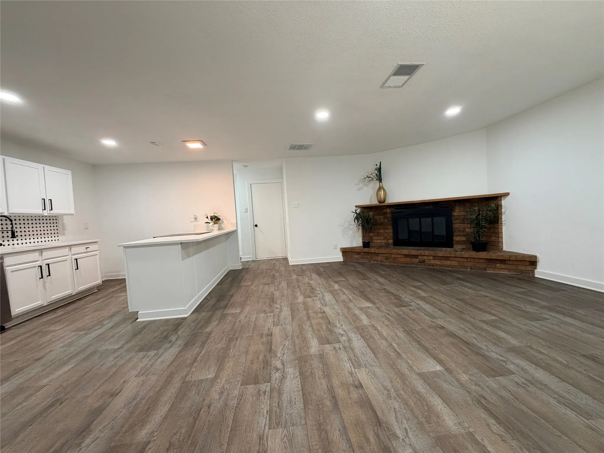 Spacious living area featuring wood-finish flooring, a built-in fireplace with a stone surround, recessed lighting, and a kitchen area with white cabinetry and a white countertop