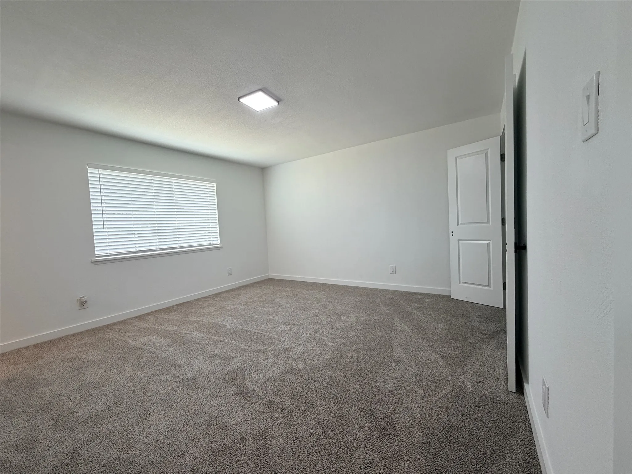 Expansive room featuring neutral-toned carpeting, a window with horizontal blinds, and a flush-mount ceiling light fixture