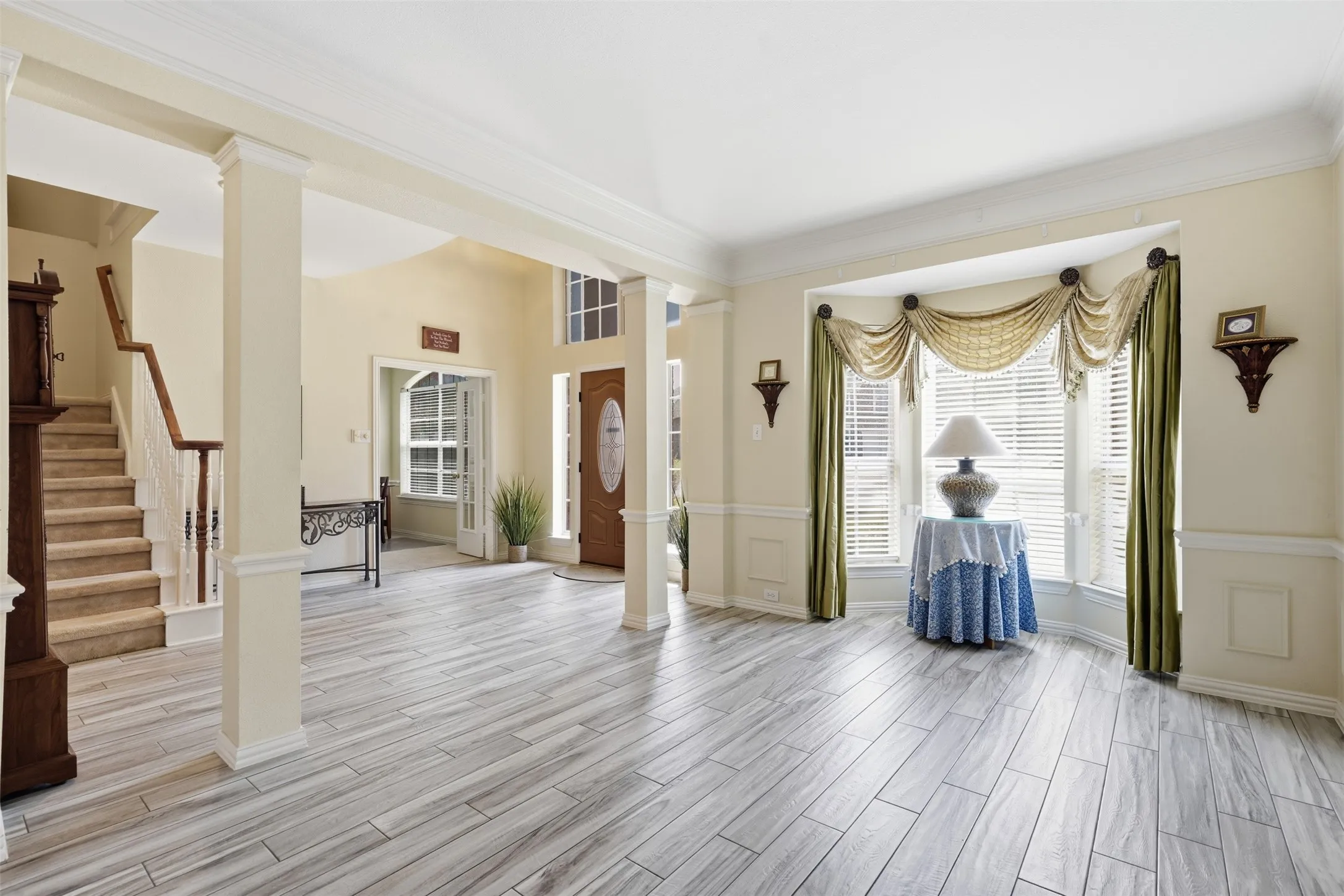 Spacious entry foyer featuring wood-finish flooring, architectural columns, a curved bay window, a staircase with a wood handrail, and a decorative front door with an oval glass insert